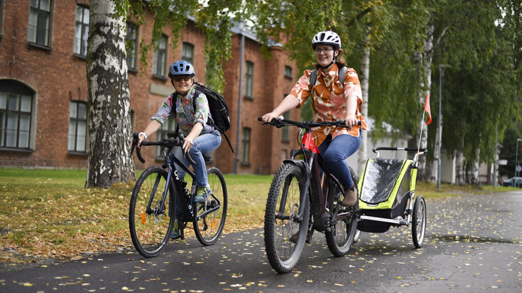 Kaksi henkilöä pyöräilee, taustalla punainen tiilirakennus ja koivuja.