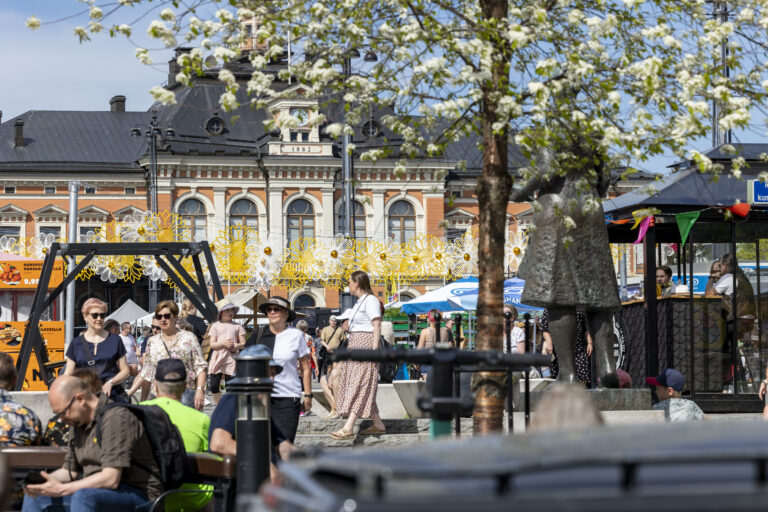 Kerroksellinen maisemakuva Kuopion torista, kuvassa etualalla ihmisiä istuskelemassa, tausta kukkaviiri ja yksi Kuopion tunnetuista maamerkeistä, Kuopion kaupungintalo.