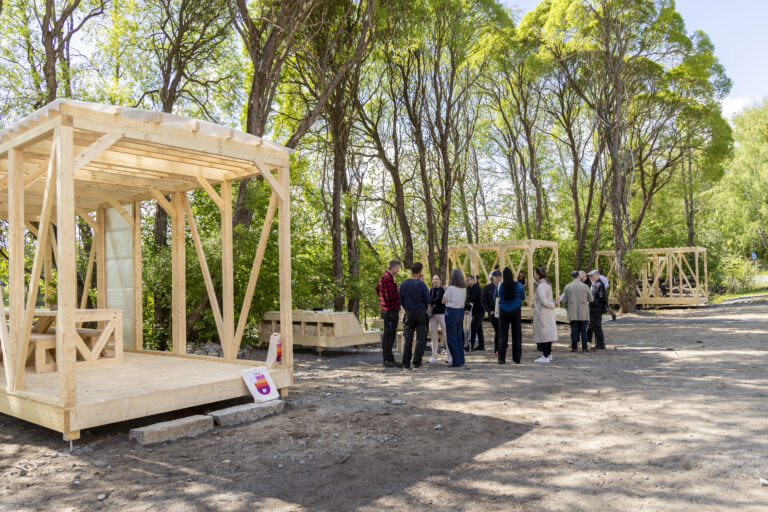 Ryhmä ihmisiä kuvan keskellä, vasemmassa reunassa puinen rakennelma, taustalla puita.