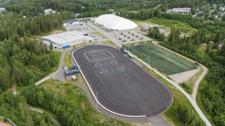 Ilmakuvassa harmaa pelikenttä, valkea kupahalli sekä metsää ja tenniskenttä.