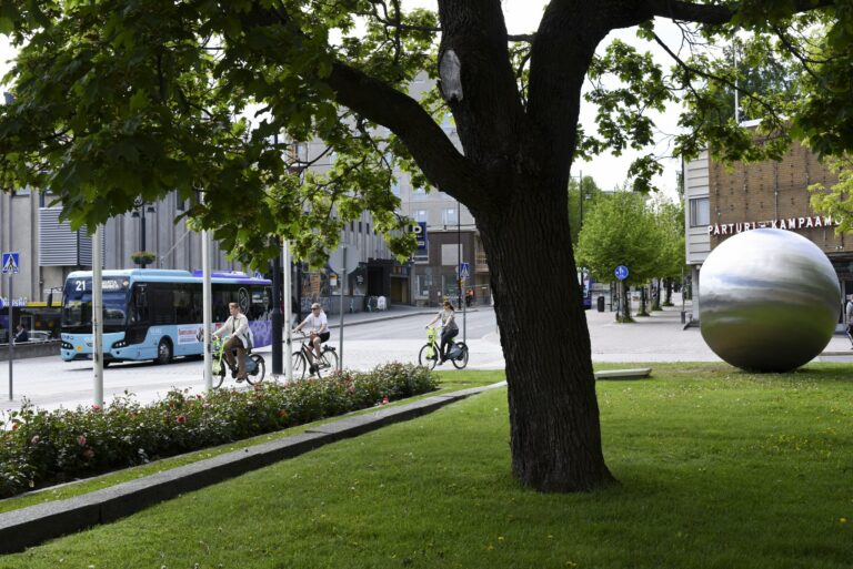 Nurmialueita, joiden keskellä puu. Taustalla katu, jossa ajaa bussi. Oikeassa reunassa metallinen pallo, joka on taideteos.
