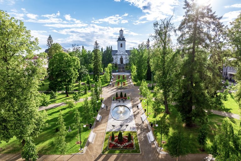 Vihreitä puita ja pensaitä kuvan molemmilla reunoilla. Keskellä taustalla vaalea tuomiokirkko sekä puiston läpi kulkeva hiekkainen kulkuväylä, jonka varrella kukkia.