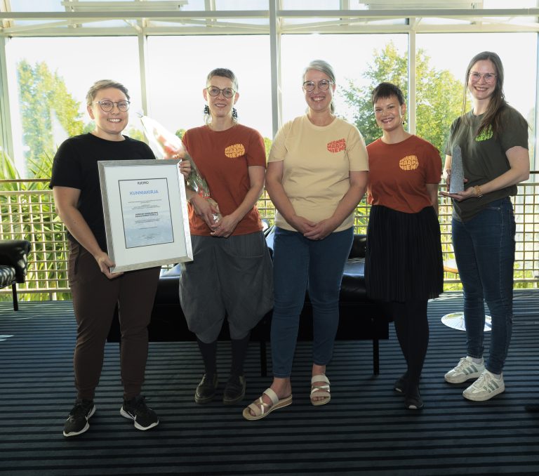 Viisi naishenkilöä seisovat rivissä ikkunoiden edessä ryhmäkuvassa.