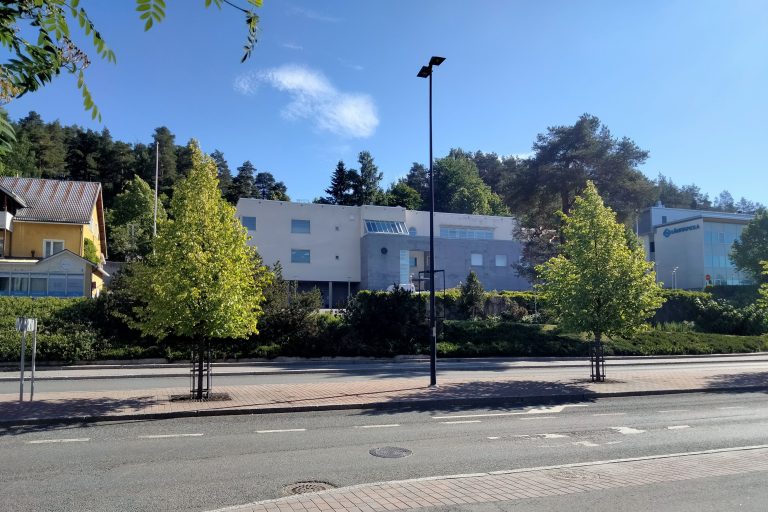 Sinistä taivasta vasten valkea rakennus, puita ja asfaltoitu katu.