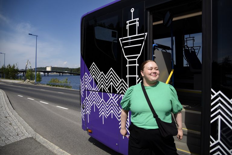 Kesäinen katu, jossa on pysähtyneenä linja-auto. Linja-autosta tulee ulos vihreäpaitainen naishenkilö.