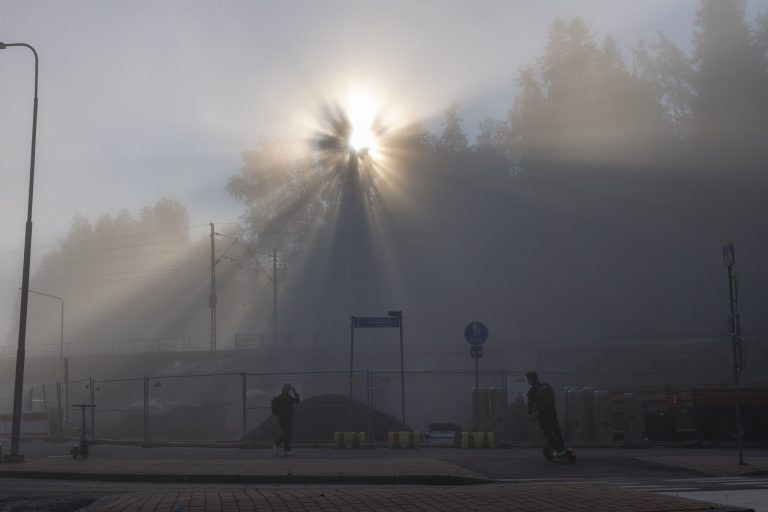 Sumuinen maisema, jossa aurinko paistaa sumun läpi luoden kauniita valonsäteitä. Taustalla on puita, ja etualalla kaksi ihmistä kävelee kadulla. Kadun varrella näkyy myös liikennemerkkejä ja aita.