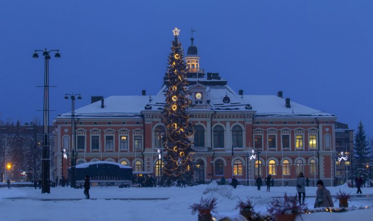 Suuri joulukuusi koristeltuna valoilla ja tähdellä latvassa seisoo lumisella torilla iltahämärässä. Taustalla näkyy näyttävä, historiallinen rakennus, jonka julkisivu on valaistu."
